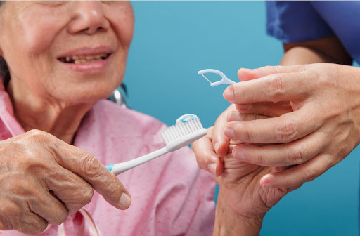 A person out of frame holds a floss pick and supports a senior woman with cleaning her teeth