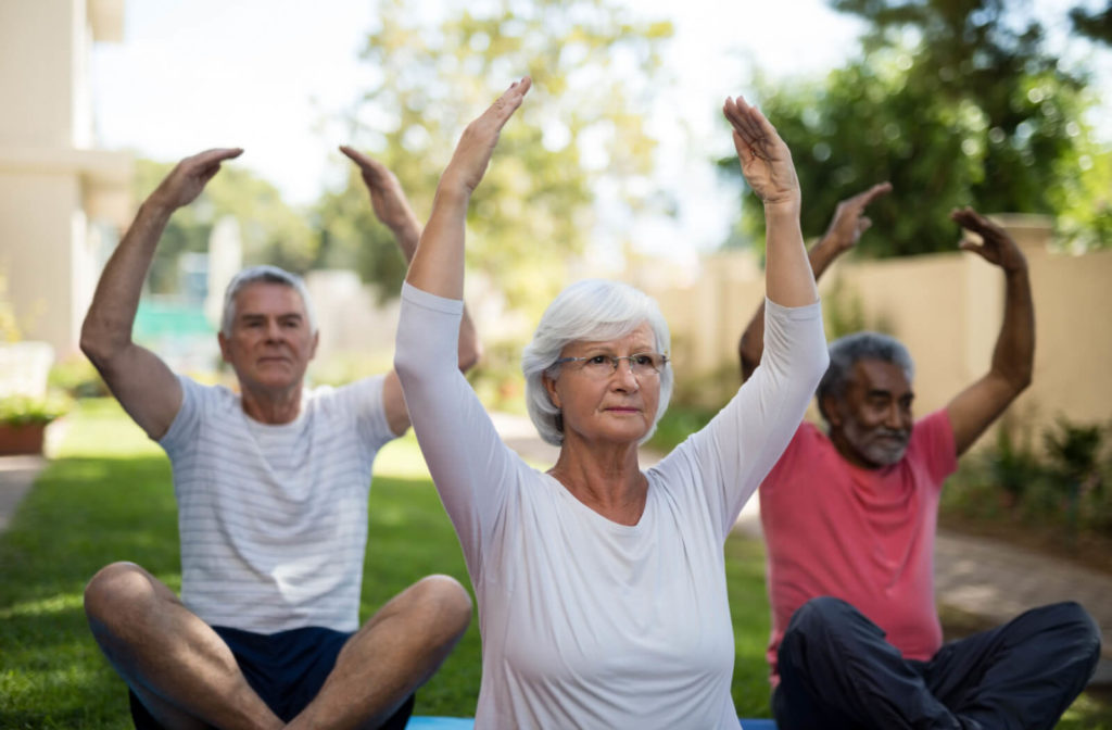 A group of seniors doing yoga outdoors with their hands raised while sitting.