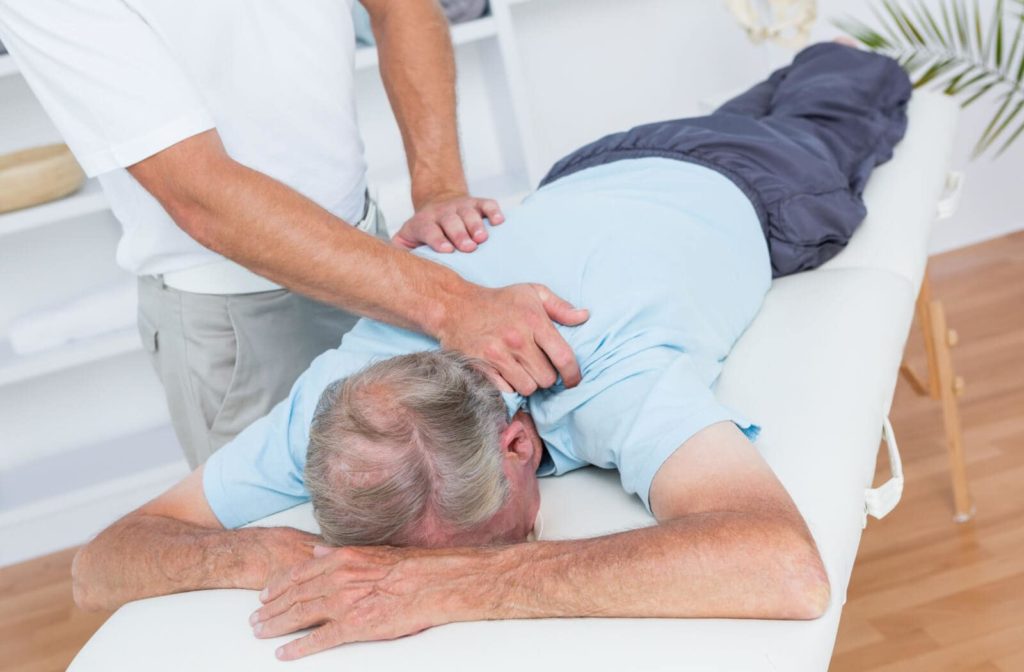A senior man lying down on a massage table while a therapist is standing behind him and giving him a massage.