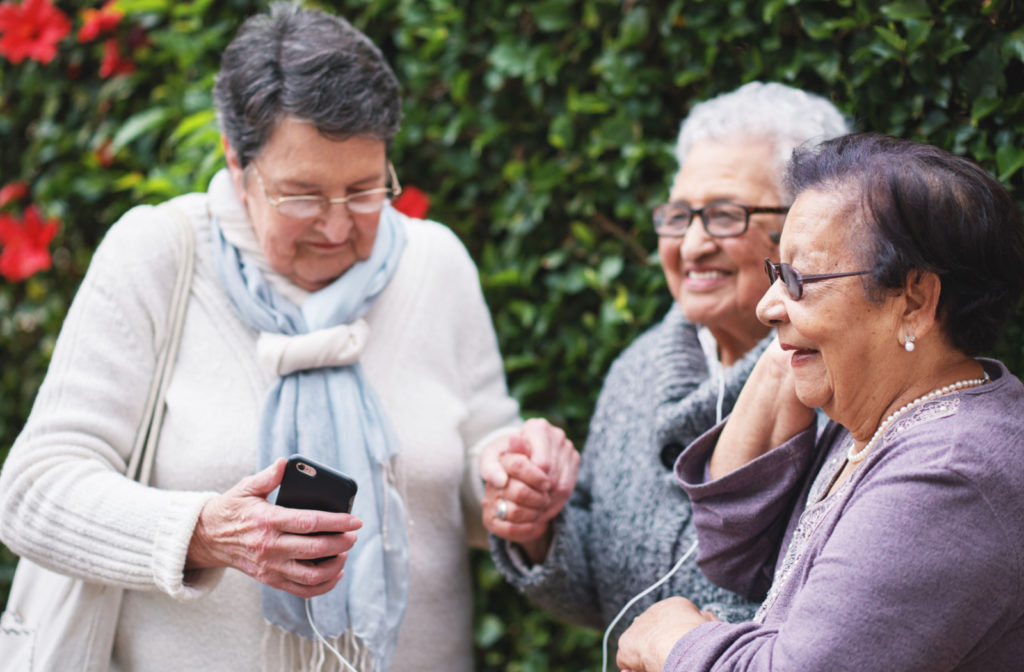 A group of senior women smiling and listening to music on a smartphone while wearing earphones