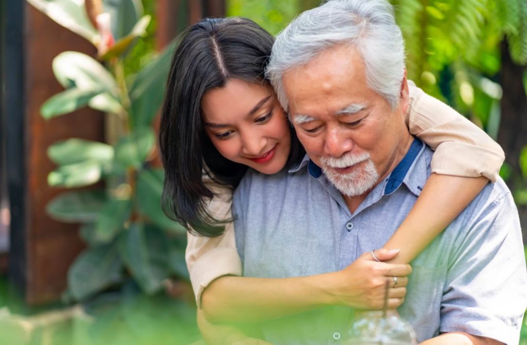 A person embraces their senior parent from behind as they both smile.