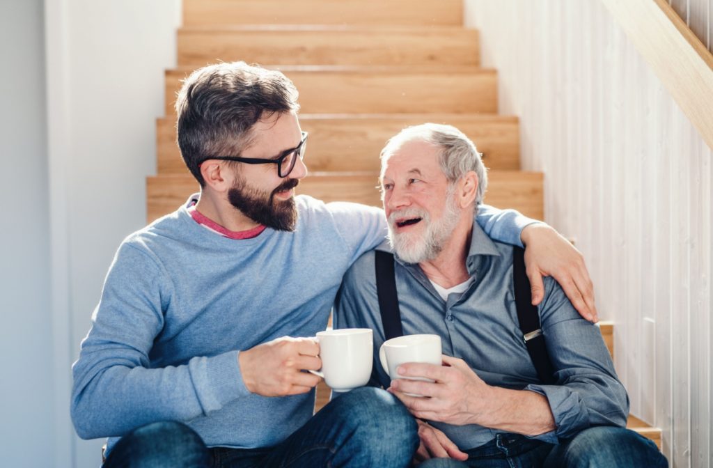 Happy senior man and adult son sitting at bottom of stairs drinking coffee