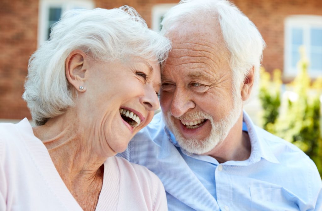 A happy older couple in assisted living sitting outdoors and smiling with their foreheads touching.