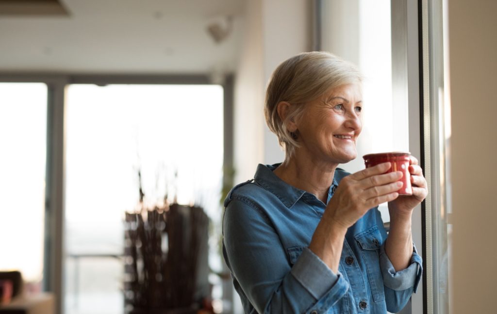 A senior sipping a red mug of coffee looks thoughtfully out of their window while soft morning light filters in.