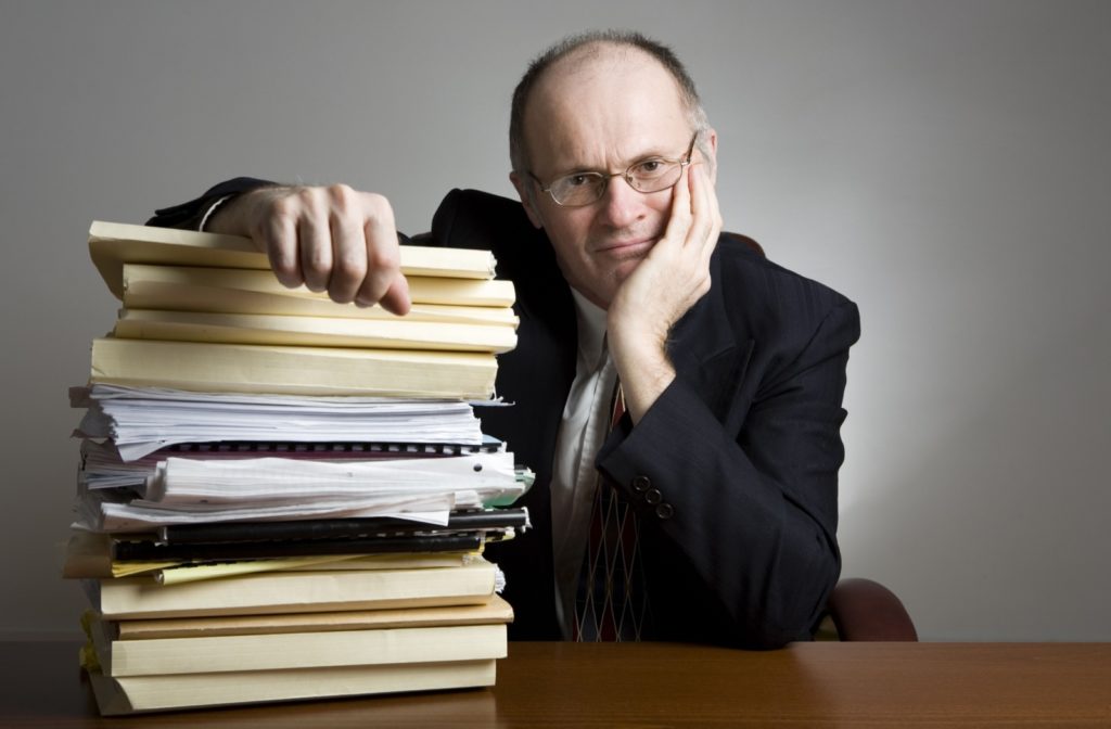 A well-dressed senior leans against a large pile of outdated paperwork, resting his head in his hand.