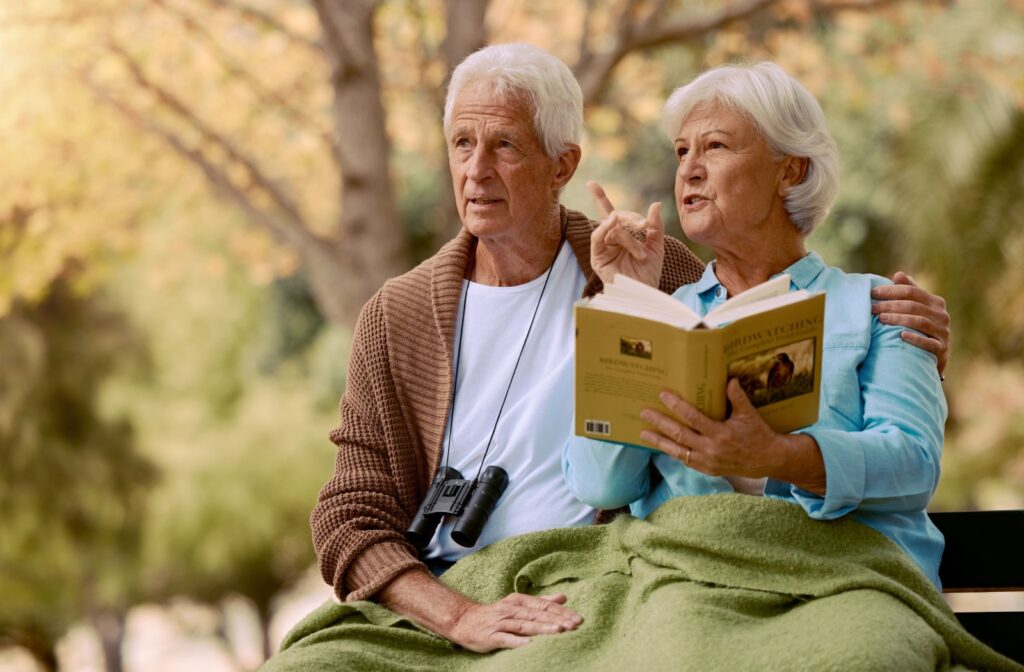 An older adult couple sits on a bench with a blanket covering them. One holds a birdwatching book while pointing at a bird off-screen, and the other has binoculars hanging around their neck.