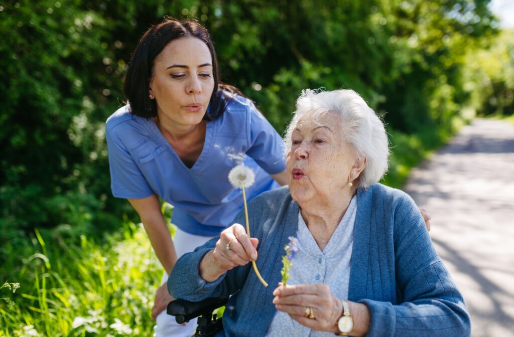 A resident in senior living outside on a walk with their caregiver, pausing to blow on a dandelion.