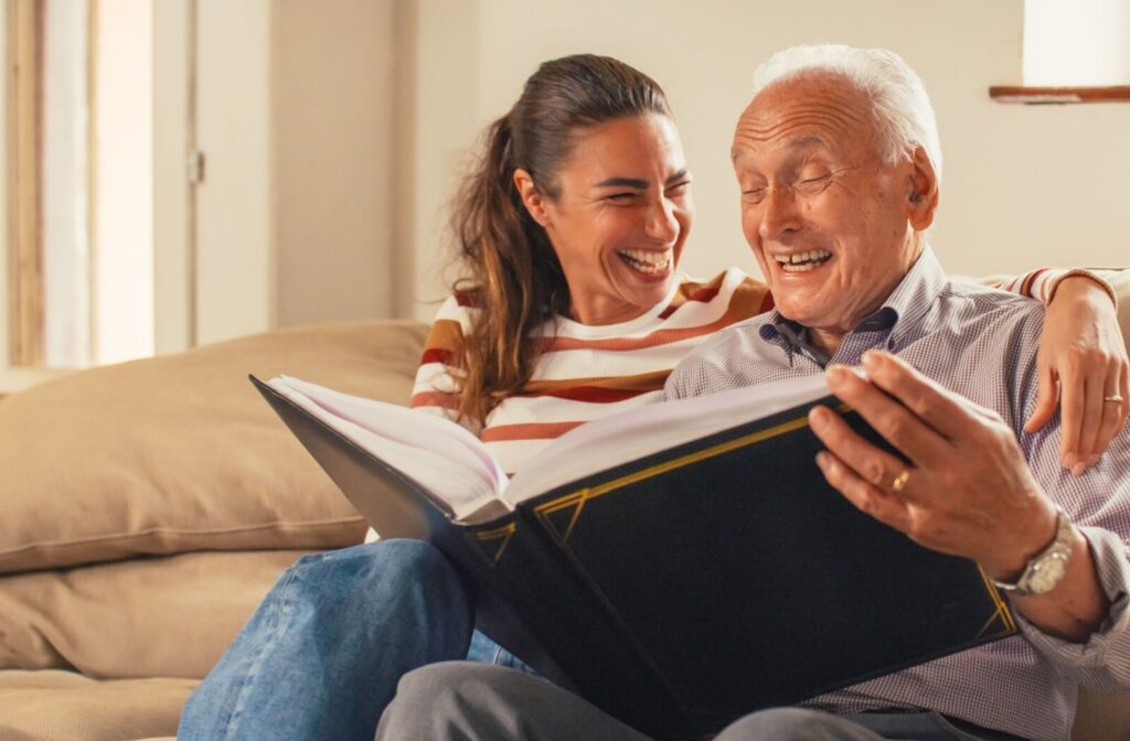 An adult child and their older parent laughing together while reminiscing with a photo album.