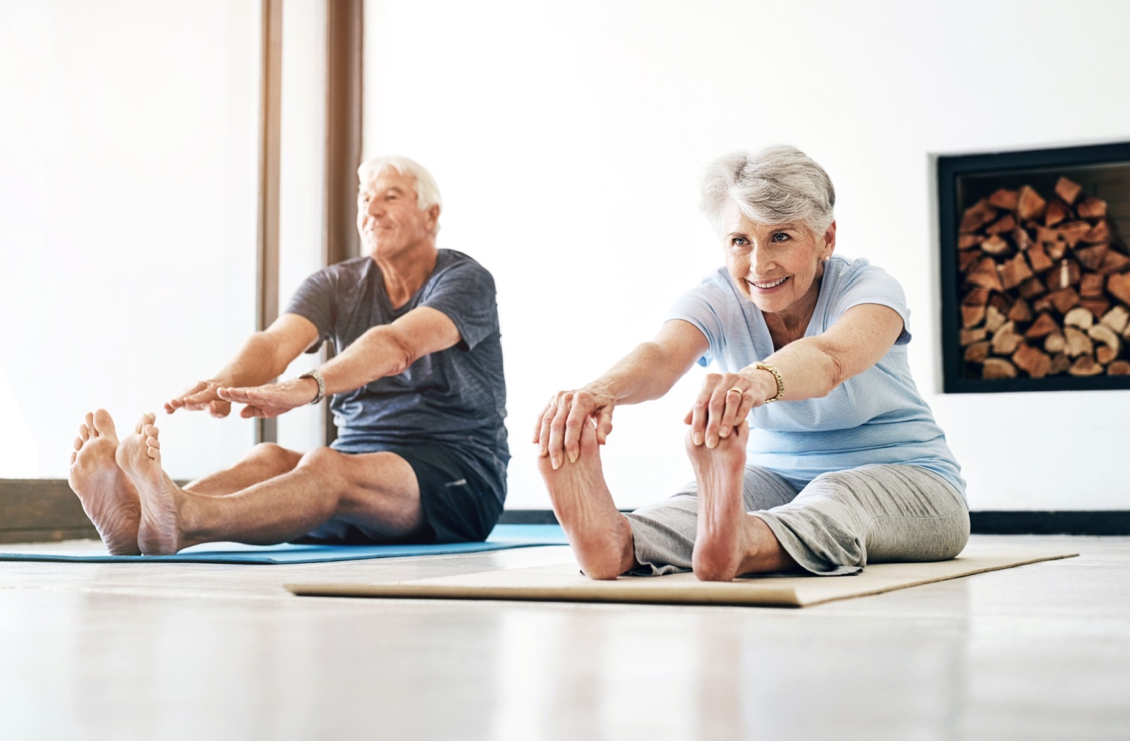 A smiling pair of older adults on yoga mats, stretching and touching their toes during an exercise class.