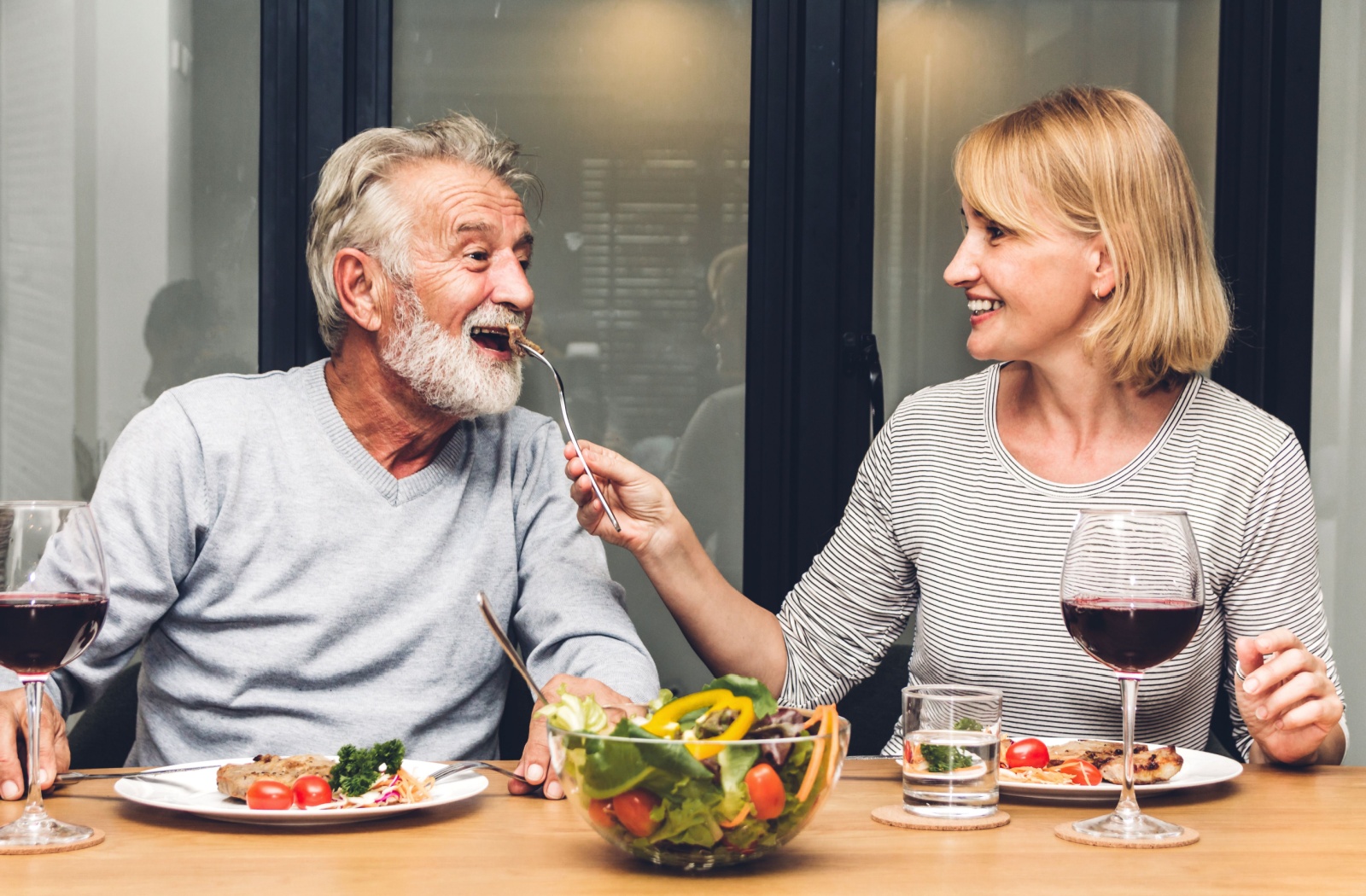 An older adult feeds their partner with dementia a bit of dinner, helping make mealtime more playful alongside colorful food