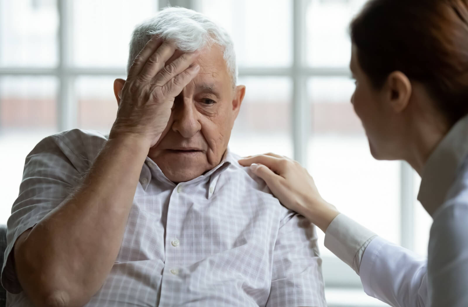 An older adult touches their forehead in confusion and asks to go home while a smiling caregiver reassures them in memory care