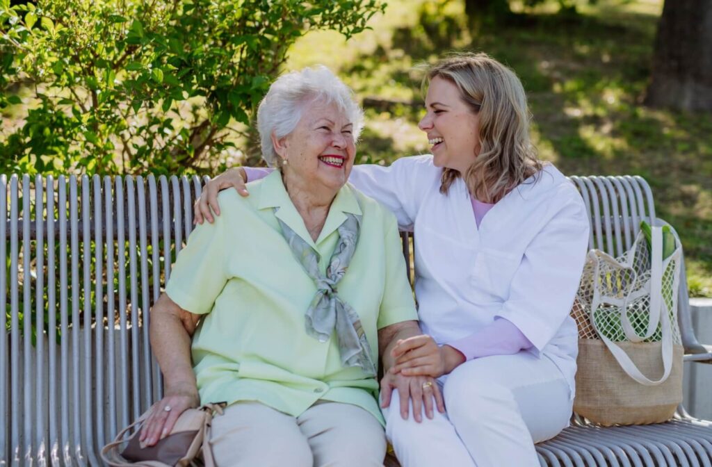 A memory care caregiver in scrubs wraps their arm around a senior while they laugh together on an outdoor bench